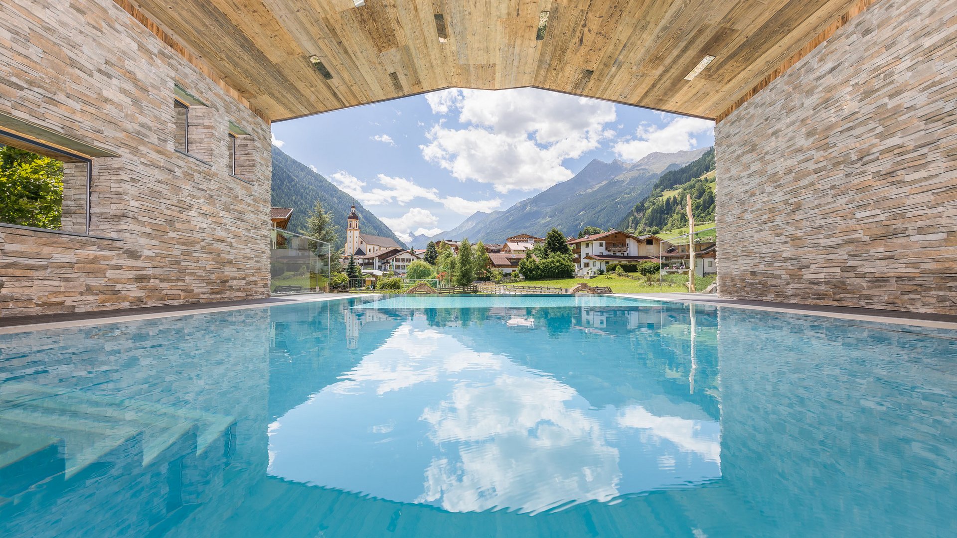 Unwind at our spa hotel in Stubaital Covered pool overlooking Alps, houses, and blue sky with clouds