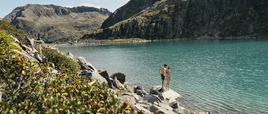 Naturliebe im Wanderhotel im Stubaital Zwei Personen am Ufer eines klaren Bergsees umgeben von Felsen und Bergen