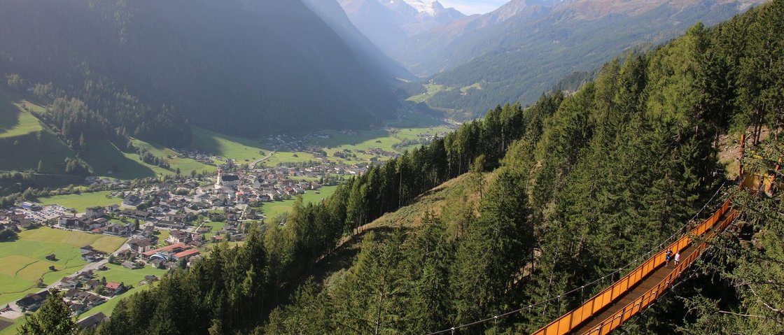 Bereit für Ihren Urlaub im Stubaital im Sommer? Hängebrücke über Wald mit Bergtal und Dorf im Hintergrund