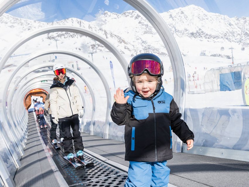 Pistenvergnügen? Ab ins Skihotel im Stubaital! Kind in Skikleidung winkt im überdachten Förderband, dahinter eine erwachsene Person und verschneite Berge